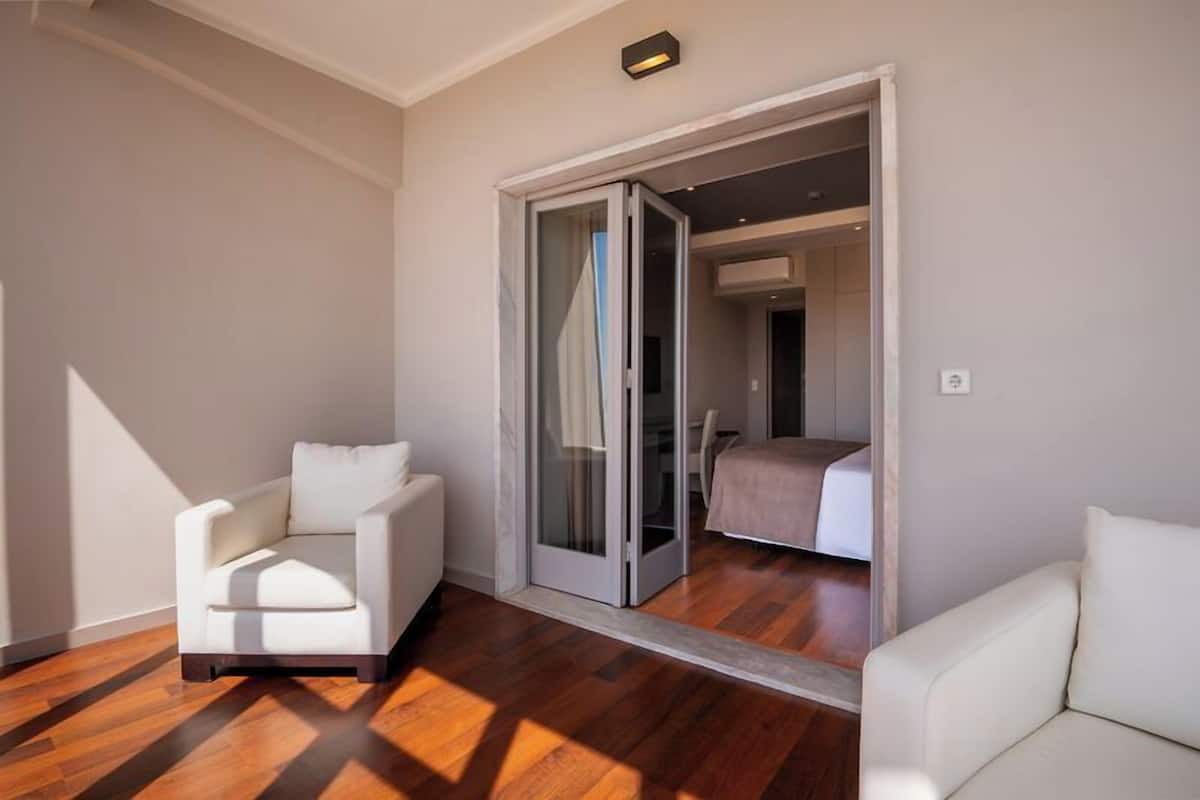 Una habitación con una silla blanca, un sofá blanco, una puerta corrediza de vidrio que da a un dormitorio y suelo de madera.