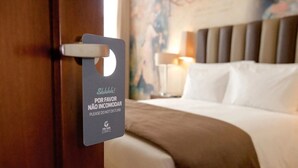 Een hotelkamer met een bed, nachtkastje en een wandlamp. Een bord op de deur vermeldt: 'Shhh! Por favor não incomodar. Please do not disturb.'