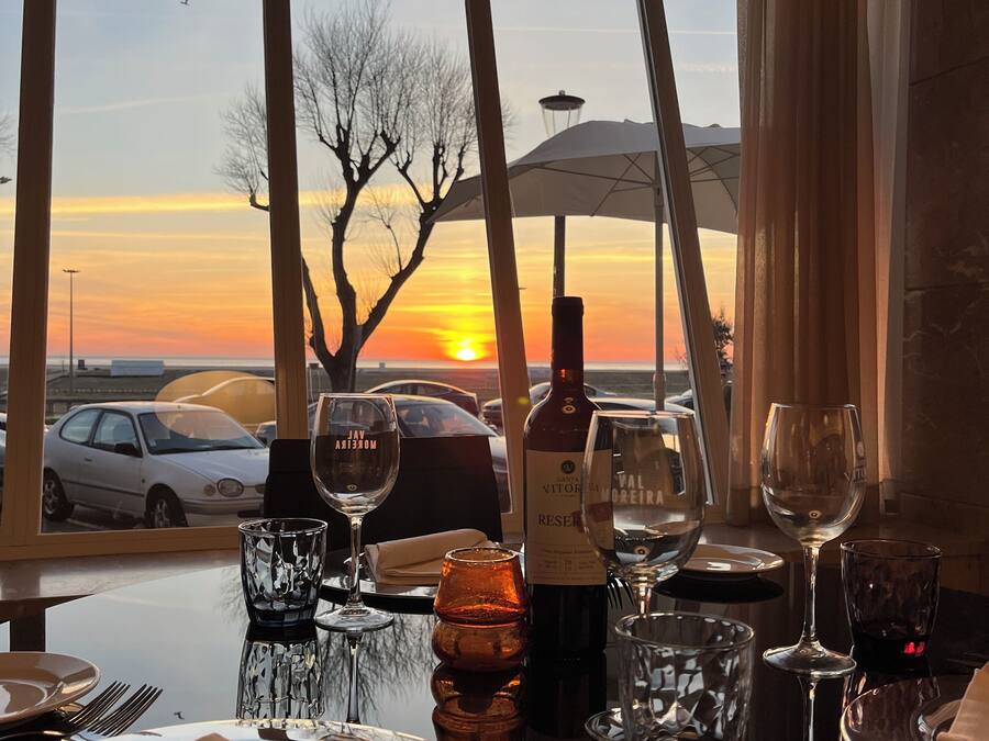 Une table dressée pour le dîner, avec une vue sur le coucher de soleil à travers de grandes fenêtres, une bouteille de vin et différents types de verres.