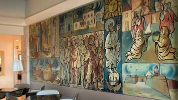 Un mural con figuras y escenas religiosas, una mesa con sillas y una lámpara en una habitación con pared lisa.