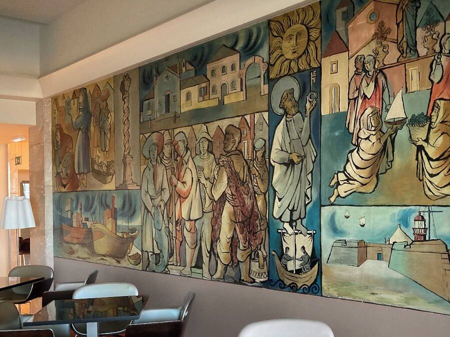 Un mural con figuras y escenas religiosas, una mesa con sillas y una lámpara en una habitación con pared lisa.