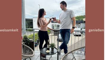 Um homem e uma mulher segurando copos de vinho em uma varanda com mesa, cadeiras e vista para um estacionamento.