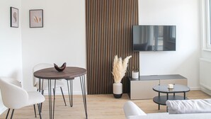 Ein modernes Wohnzimmer mit einer Couch, zwei Sesseln, einem runden Tisch, einem kleinen Beistelltisch, einem an der Wand montierten Fernseher und einer Zierpflanze.