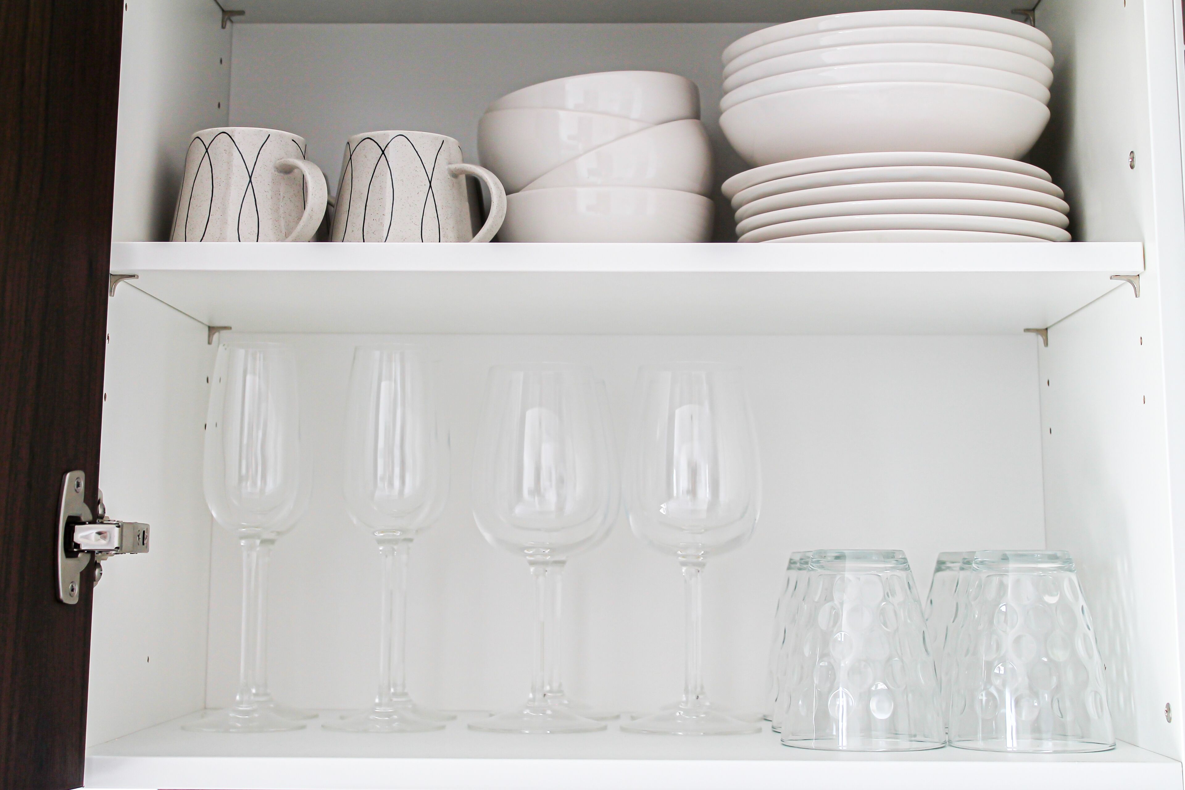 Un armario blanco con estantes que contienen tazas blancas, platos y vasos de vidrio transparente.