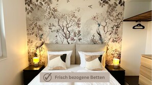 Ein Schlafzimmer mit floralem Wandpapier, zwei Nachttischlampen, einer Holzkommode und einem Bett mit zwei Kissen und zwei Handtüchern.