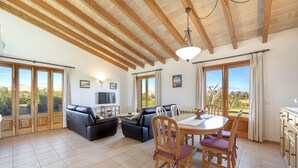 A spacious living room with a dining area, wooden ceiling beams, large windows, and a view of the outdoors. - Els Evols for 6 with pool between Pollensa and Alcudia (Alcúdia)