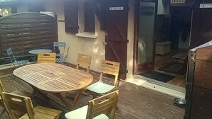 Een terras van hout buiten, met een ronde tafel en stoelen, een schuifdeur naar een overdekt gedeelte en een kleine buitenkeuken.