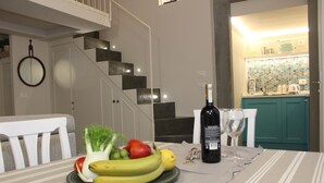 Ein Esstisch mit Obstschale, Weinflasche und Gläsern. Im Hintergrund sind eine Treppe und eine Küche zu sehen.