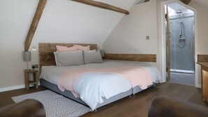 Un dormitorio con techo inclinado, una cama con cabecera de madera, una mesita de noche con lámpara y una zona de ducha visible a través de una puerta abierta.