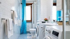 Une petite buanderie comprenant un lave-linge et un sèche-linge, une fenêtre avec un rideau bleu et un radiateur fixé au mur.