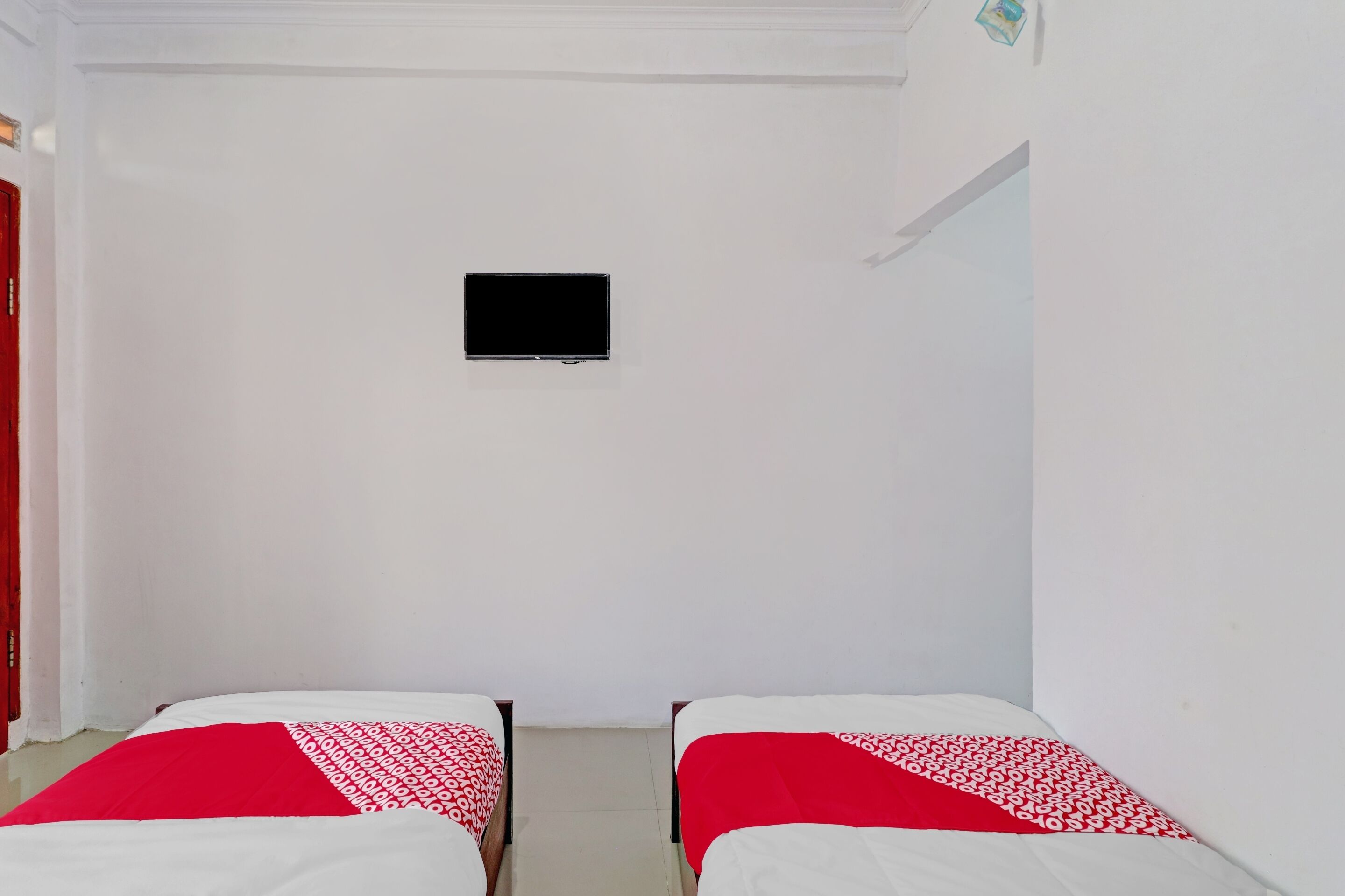 Una habitación con dos camas, cada una con una colcha de cuadros rojos y blancos, un televisor de pantalla plana montado en la pared y paredes blancas.