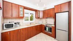 A modern kitchen with wooden cabinets, a stainless steel refrigerator, a microwave, a kettle, and a window with a view of greenery. - Europe Villa Io - Two Bedroom Villa, Sleeps 4 (Ionian Islands)