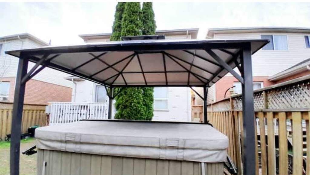 A covered outdoor seating area with a cushioned bench, surrounded by a wooden fence and a tall tree.