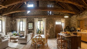 Un intérieur rustique avec des murs en pierre, des poutres apparentes, un coin repas avec une table et des chaises, une cuisine avec des meubles en bois et un lustre.