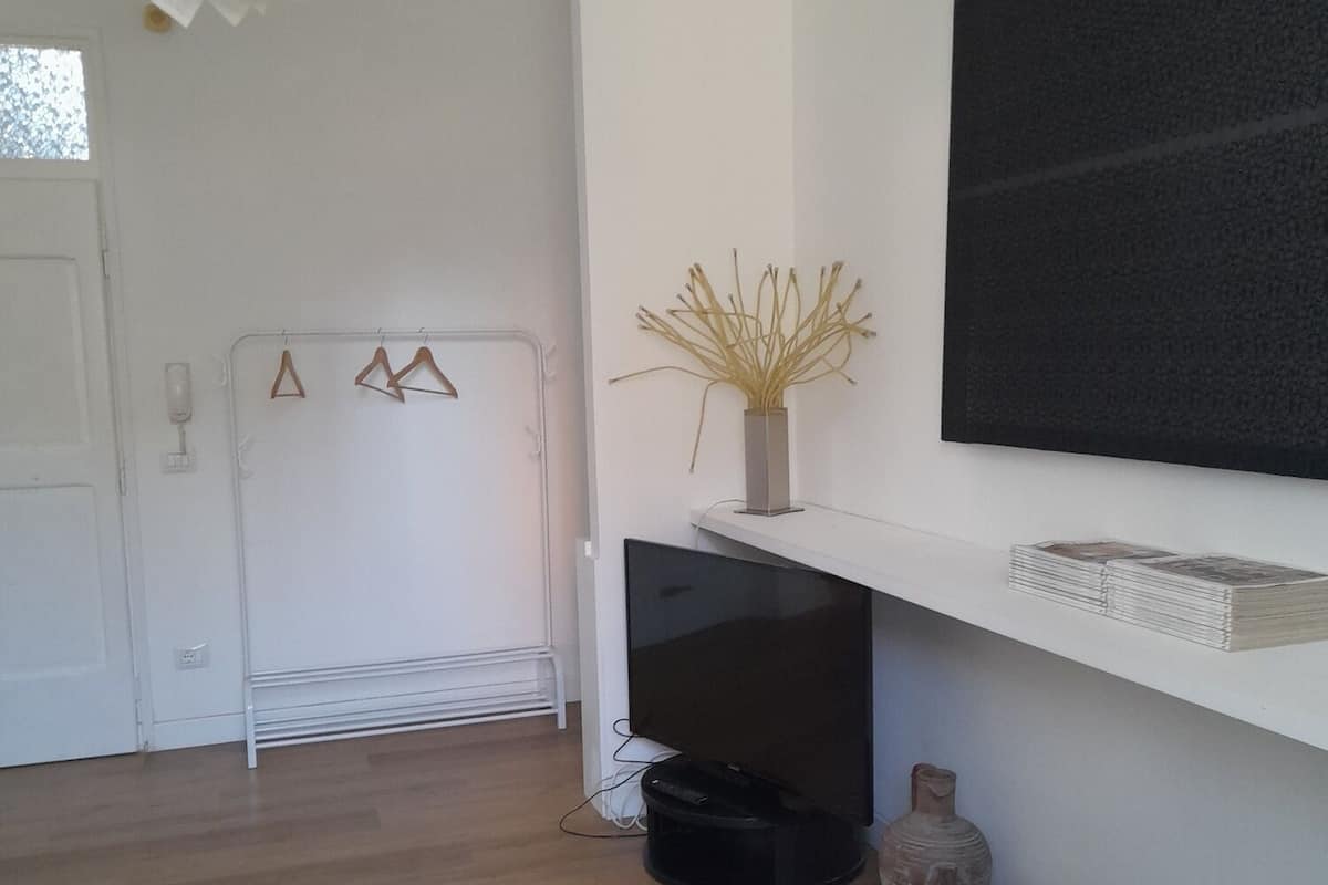 Un salón minimalista con pared blanca, un televisor montado, una estantería blanca con libros, un radiador y una lámpara colgante.