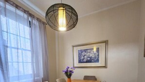 Un comedor con mesa de madera, sillas grises, lámpara colgante, un cuadro en la pared y una ventana con cortinas translúcidas.