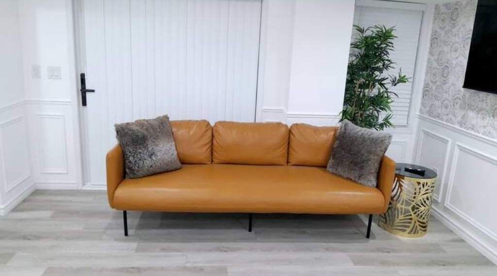 A tan leather sofa with two pillows, positioned in a room with white paneled walls and a wooden floor.