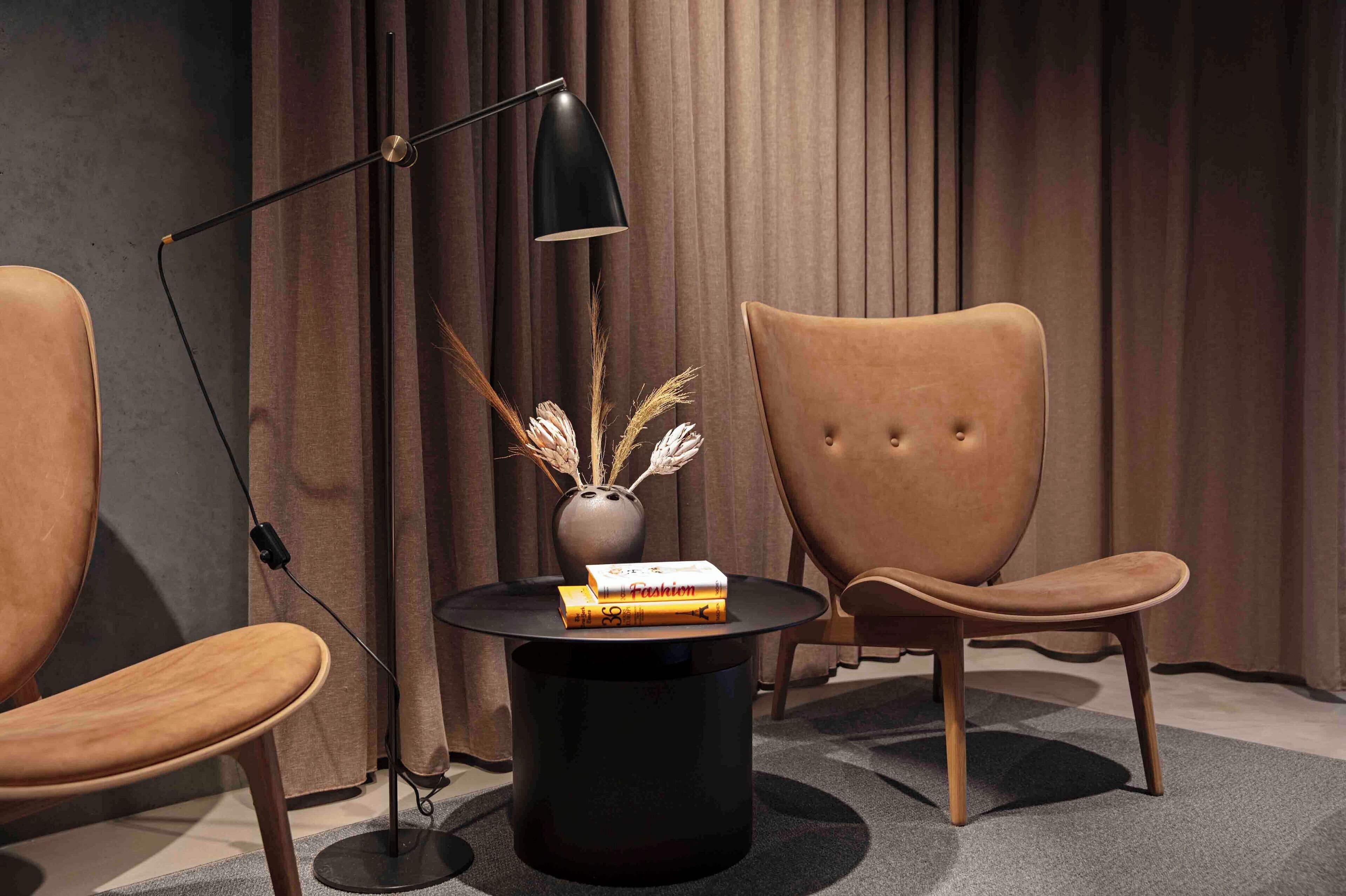 Een moderne kamer met twee leren fauteuils, een zwarte vloerlamp, een vaas met gedroogde planten en een stapel tijdschriften op een zwart nachtkastje.