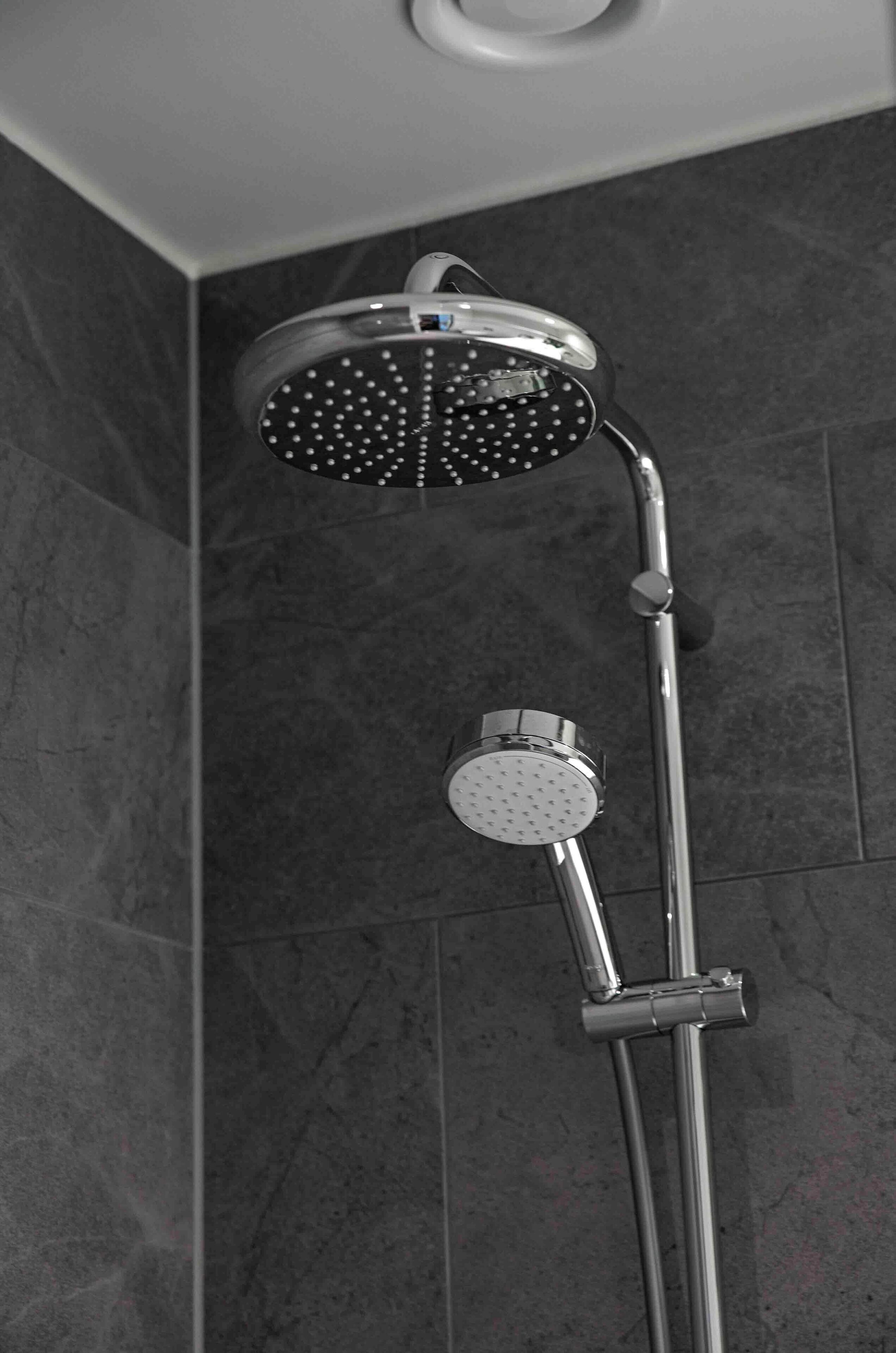 Ducha moderna con cabezal de ducha de lluvia y cabezal de ducha manual, montados en una estructura de cromo frente a una pared de azulejos.