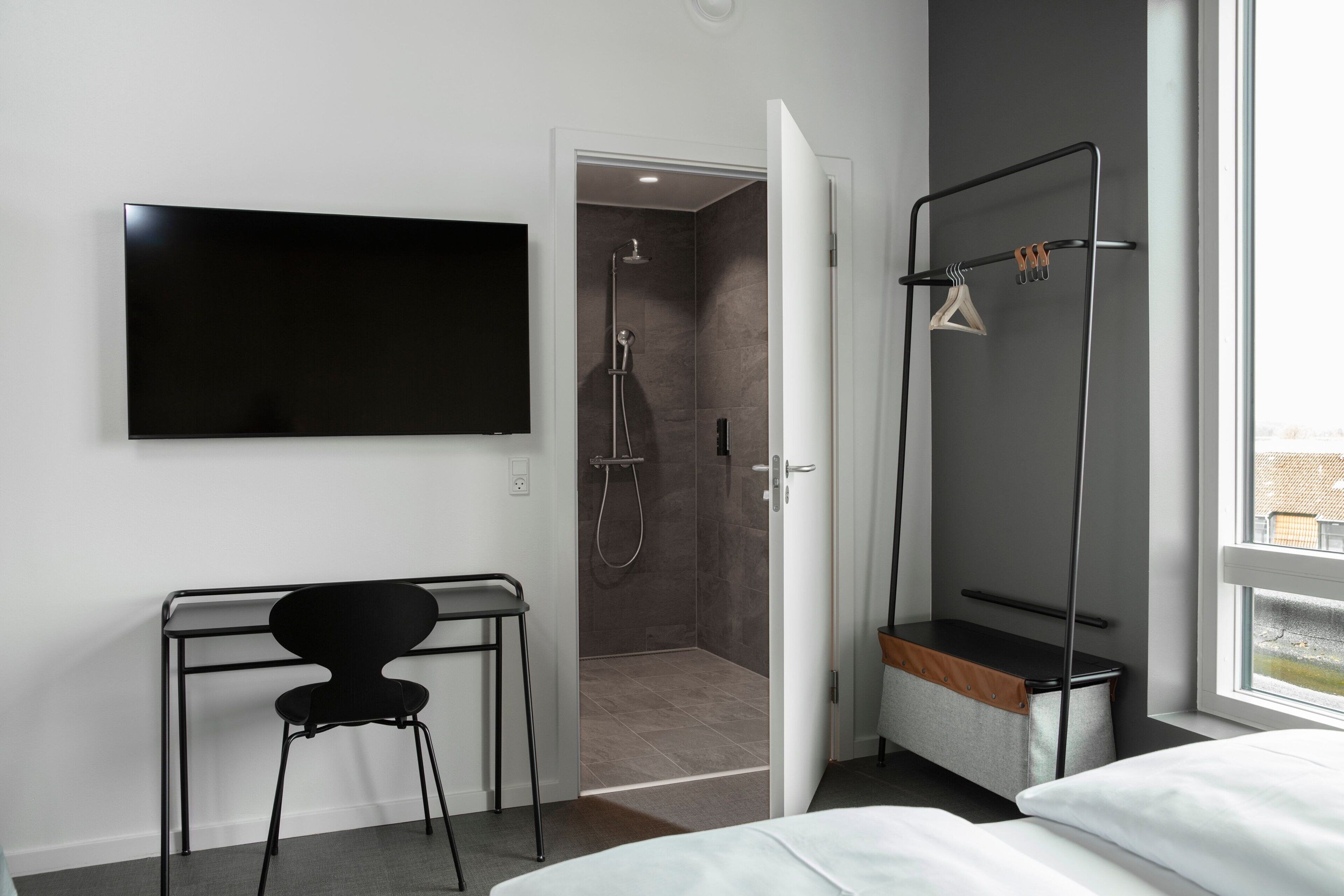 A modern hotel room with a bed, a desk with a chair, a TV mounted on the wall, a shower area, and a window with a view.