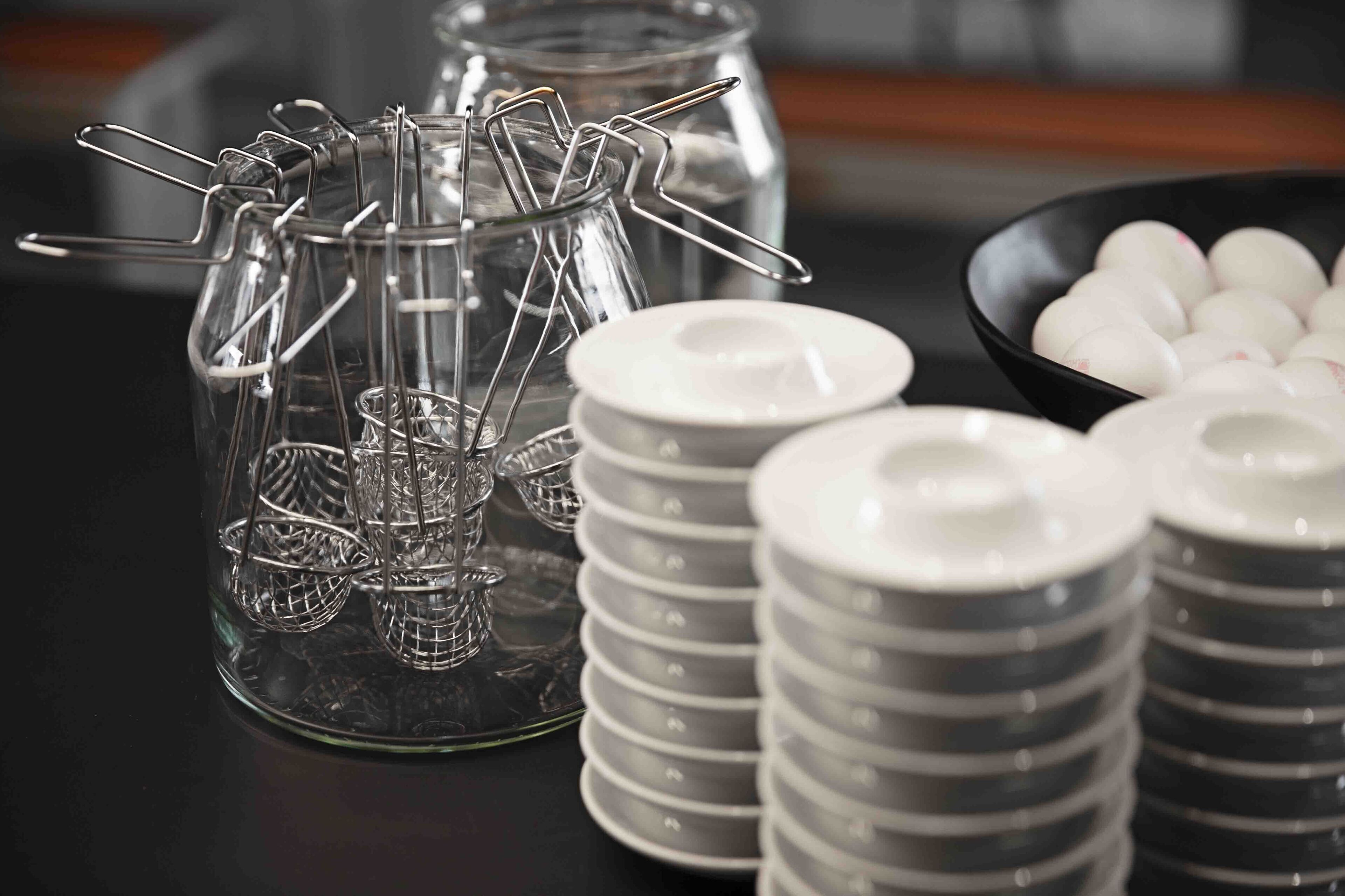 Een glazen pot met metalen draadbaskets, gestapelde grijze en witte borden, en een schaal met witte, bolvormige objecten.