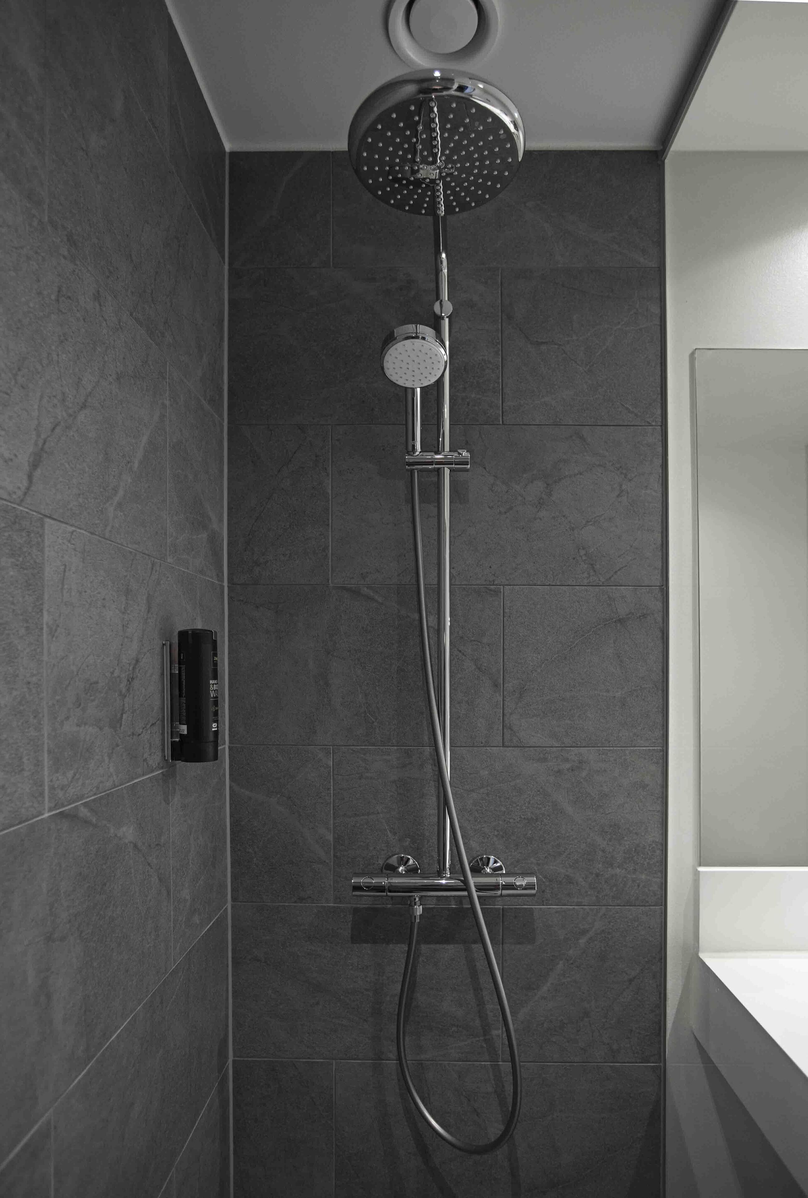 Une douche moderne dotée d’un pommeau de douche à main et d’une douchette à pluie fixée au mur, le tout installé sur un mur recouvert de carreaux foncés.