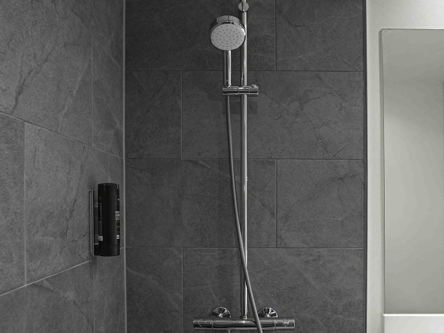 Une douche moderne dotée d’un pommeau de douche à main et d’une douchette à pluie fixée au mur, le tout installé sur un mur recouvert de carreaux foncés.