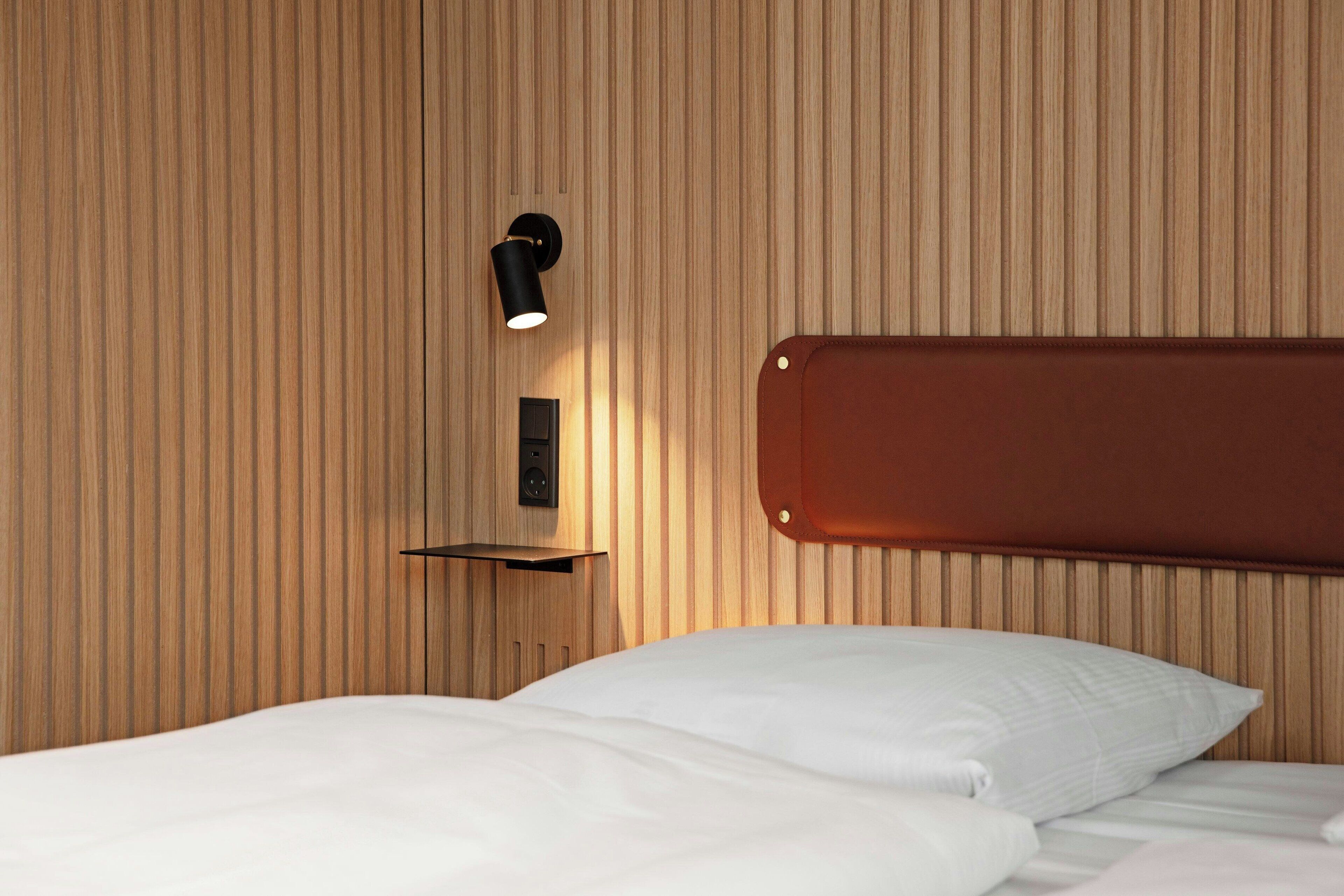 Una cama bien hecha con sábanas blancas, un estante de pared con luz negra y un panel de pared de madera.