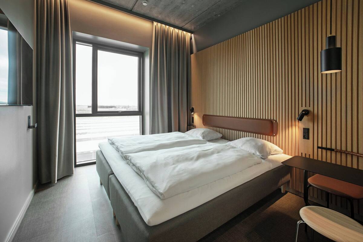 Quarto de hotel moderno com duas camas, uma janela grande e vista para a cidade.
