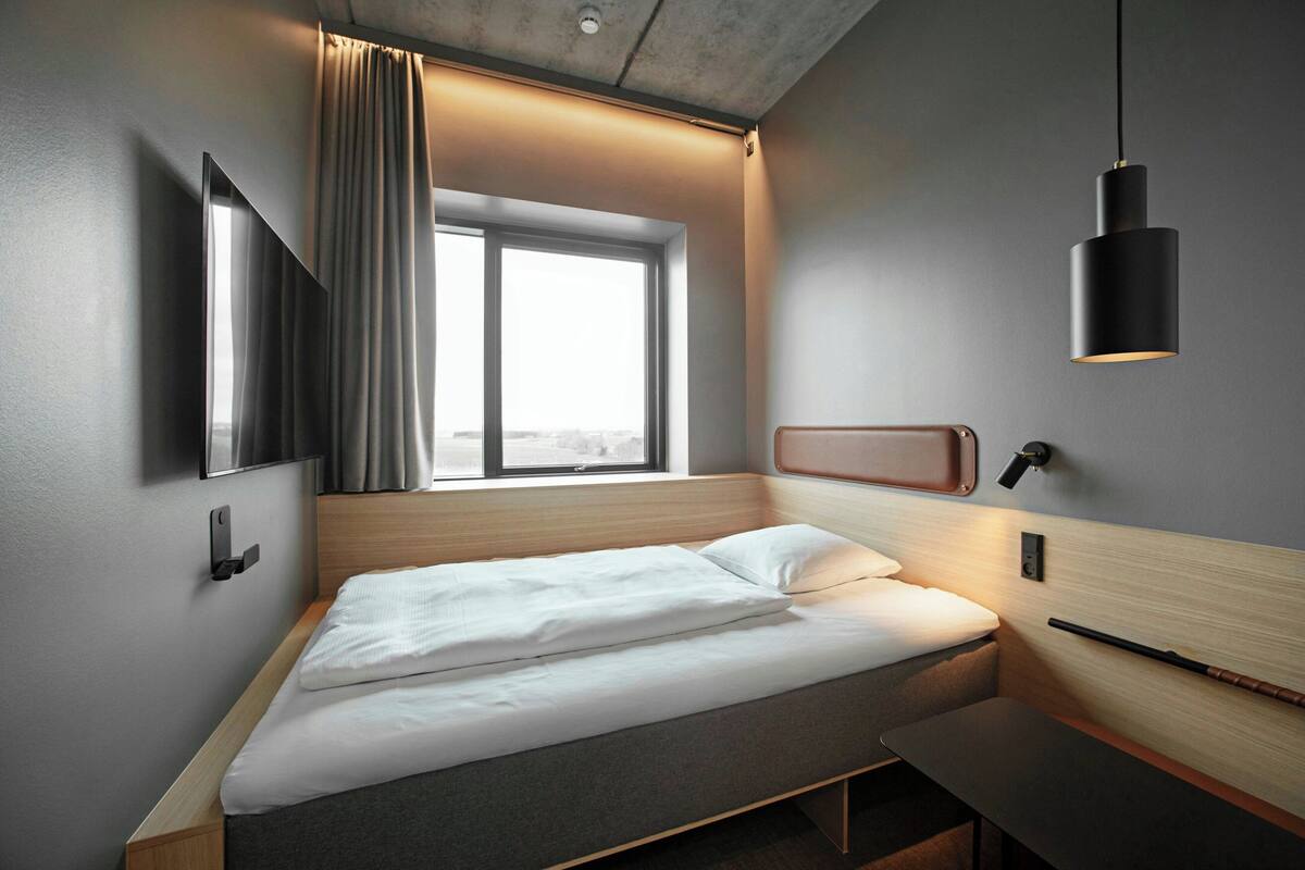 Quarto de hotel moderno com cama, janela com cortinas, televisão fixada na parede e luminária pendente.