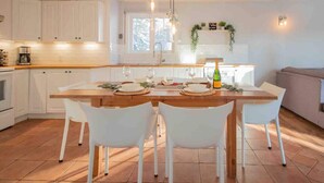 A modern kitchen with white cabinets, a wooden dining table, and white chairs. There is a window providing natural light and a hanging light fixture above the table.
