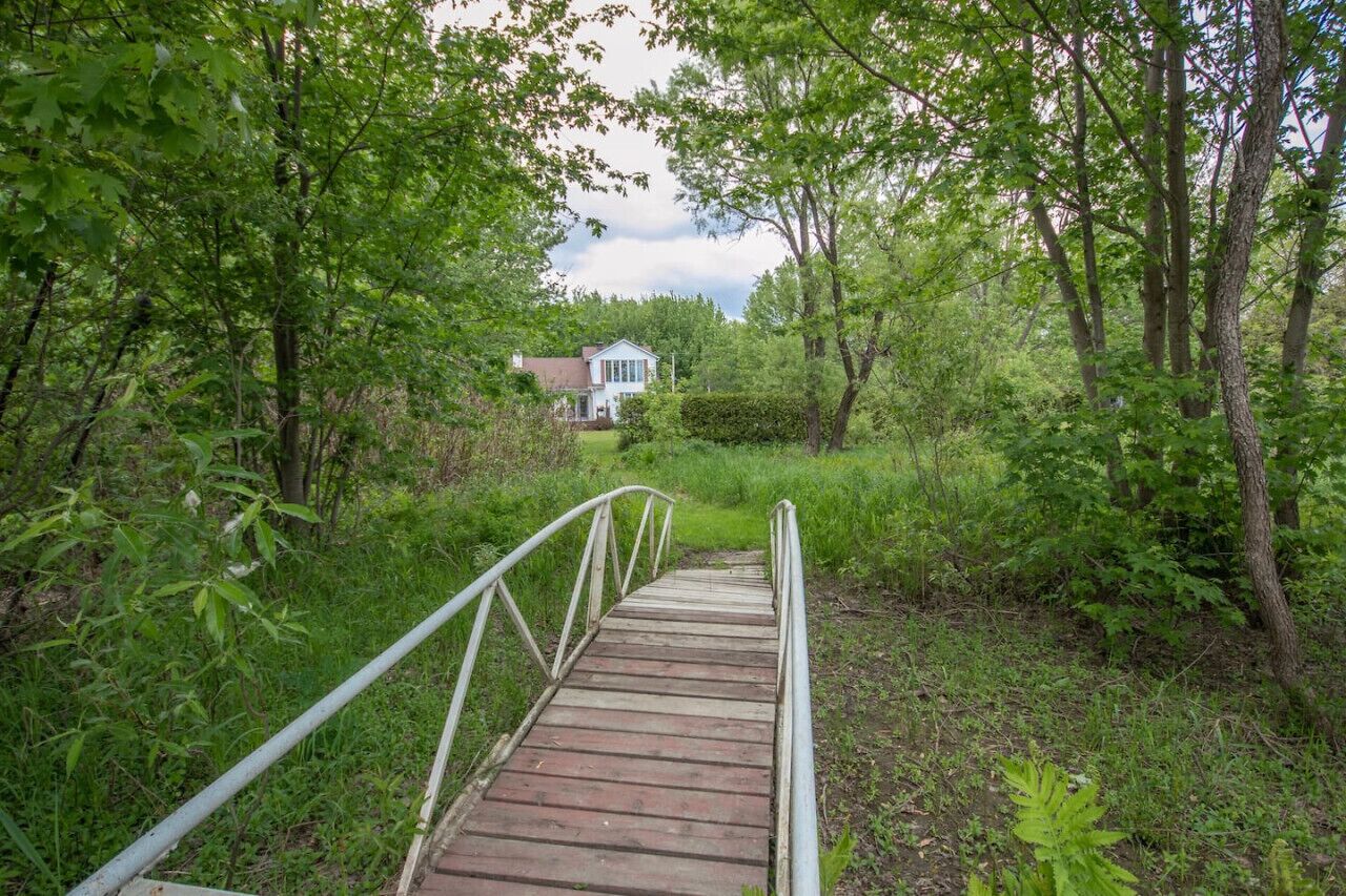 A wooden bridge leading to a house surrounded by dense greenery.