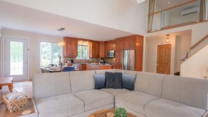 A modern kitchen with wooden cabinets, a stainless steel refrigerator, and a dining area with a light-colored sofa and a woven coffee table.