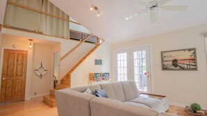 A spacious living room with a high ceiling, a staircase, a large window, and a painting on the wall.