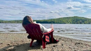 A person sitting on a red chair by a lake with a drink in hand, overlooking a forested hill.