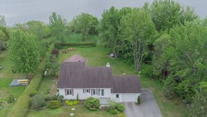 A white house with a dark roof, surrounded by lush greenery and a lake.