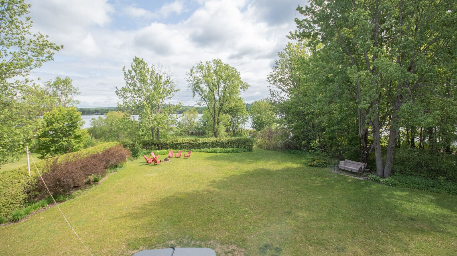 A grassy outdoor area with a bench and red chairs, surrounded by trees and a hedge.