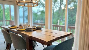 A wooden dining table with chairs, a chandelier, and a view of greenery through large windows. - Chalet BIA - Au courant des 2 rivières (Weedon)