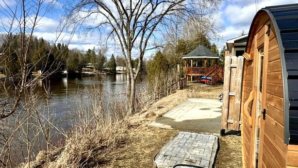 A wooden cabin with a curved roof by a lake, a concrete pathway leading to it, and a small wooden structure in the background. - Chalet BIA - Au courant des 2 rivières (Weedon)