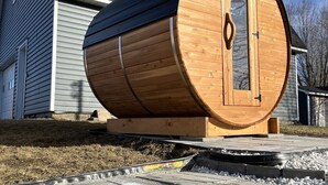 A large wooden barrel structure with a door, placed on a paved area in front of a house. - Chalet BIA - Au courant des 2 rivières (Weedon)
