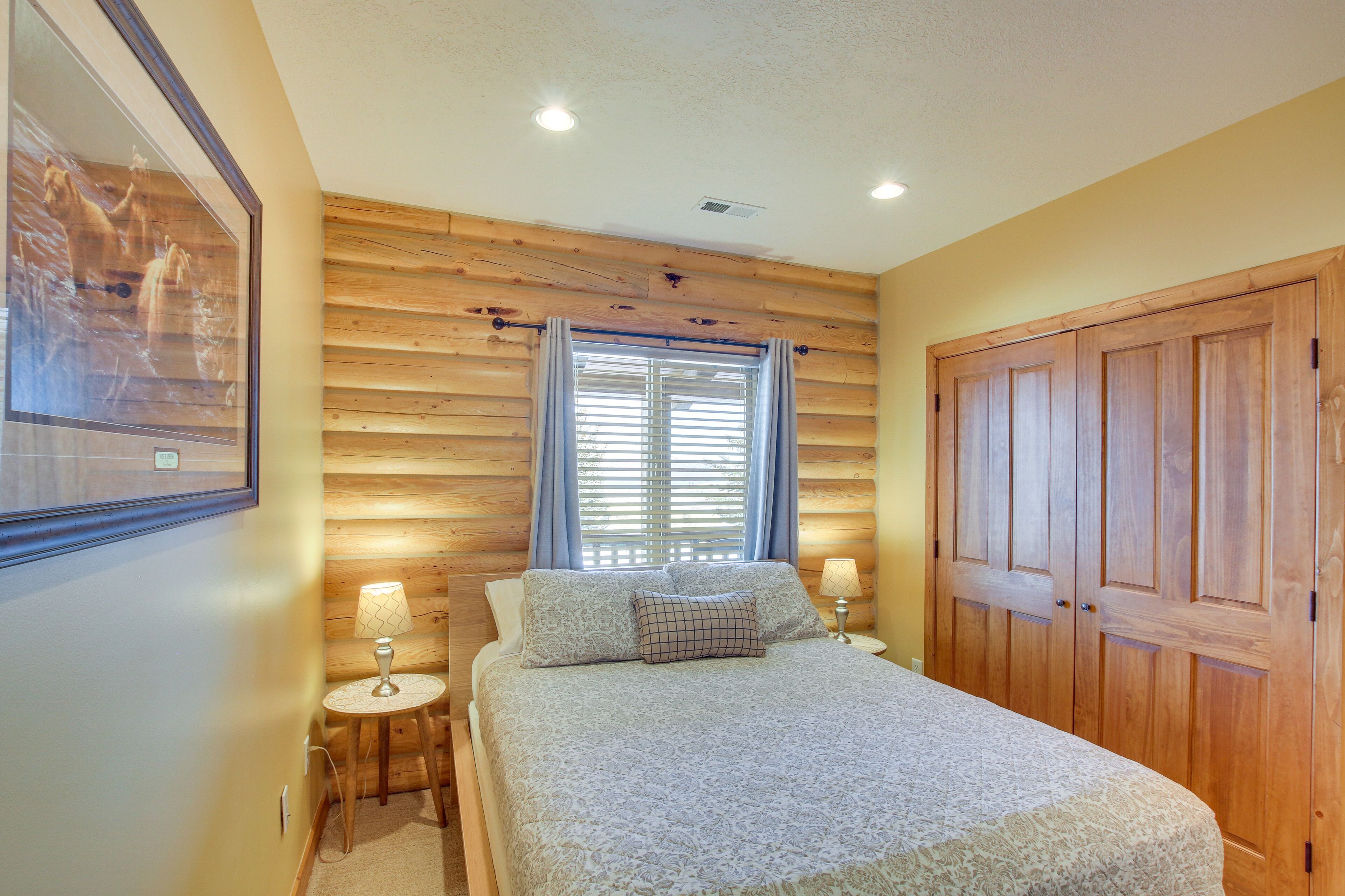 A bedroom with a bed, a nightstand, a lamp, a window with blinds, and a wooden wardrobe.