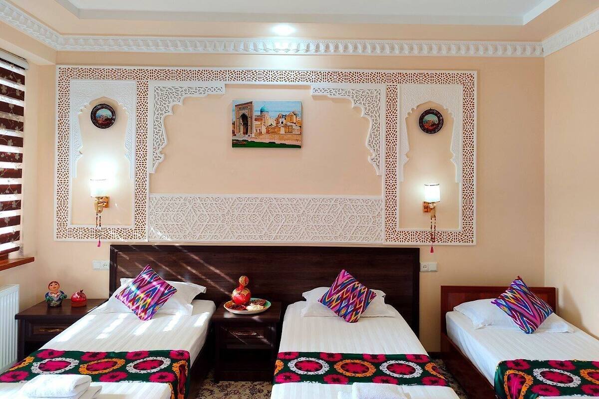 Ein Hotelzimmer mit drei Betten, jedes mit farbenfroher Bettwäsche und einem dekorativen Kopfteil. An den Wänden sind Lampen angebracht und ein Gemälde hängt über den Betten.