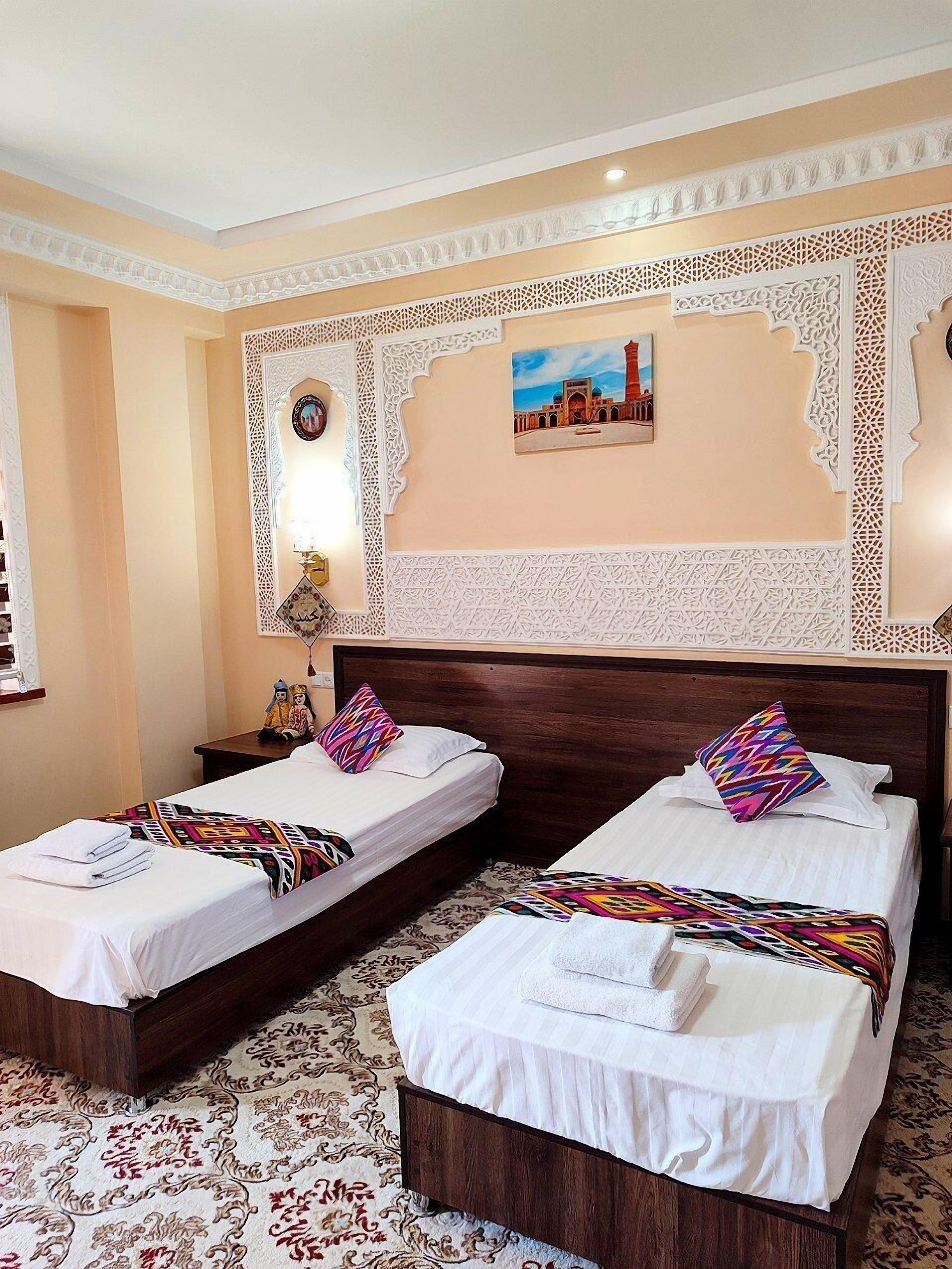 A hotel room with two beds, each with a patterned bedspread and pillows, a decorative wall with intricate designs, and a framed picture of a building.