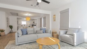 A spacious living room with a grey sofa, a wooden coffee table, and a grey armchair. There is a ceiling fan, a kitchen in the background, and a large window with blinds.