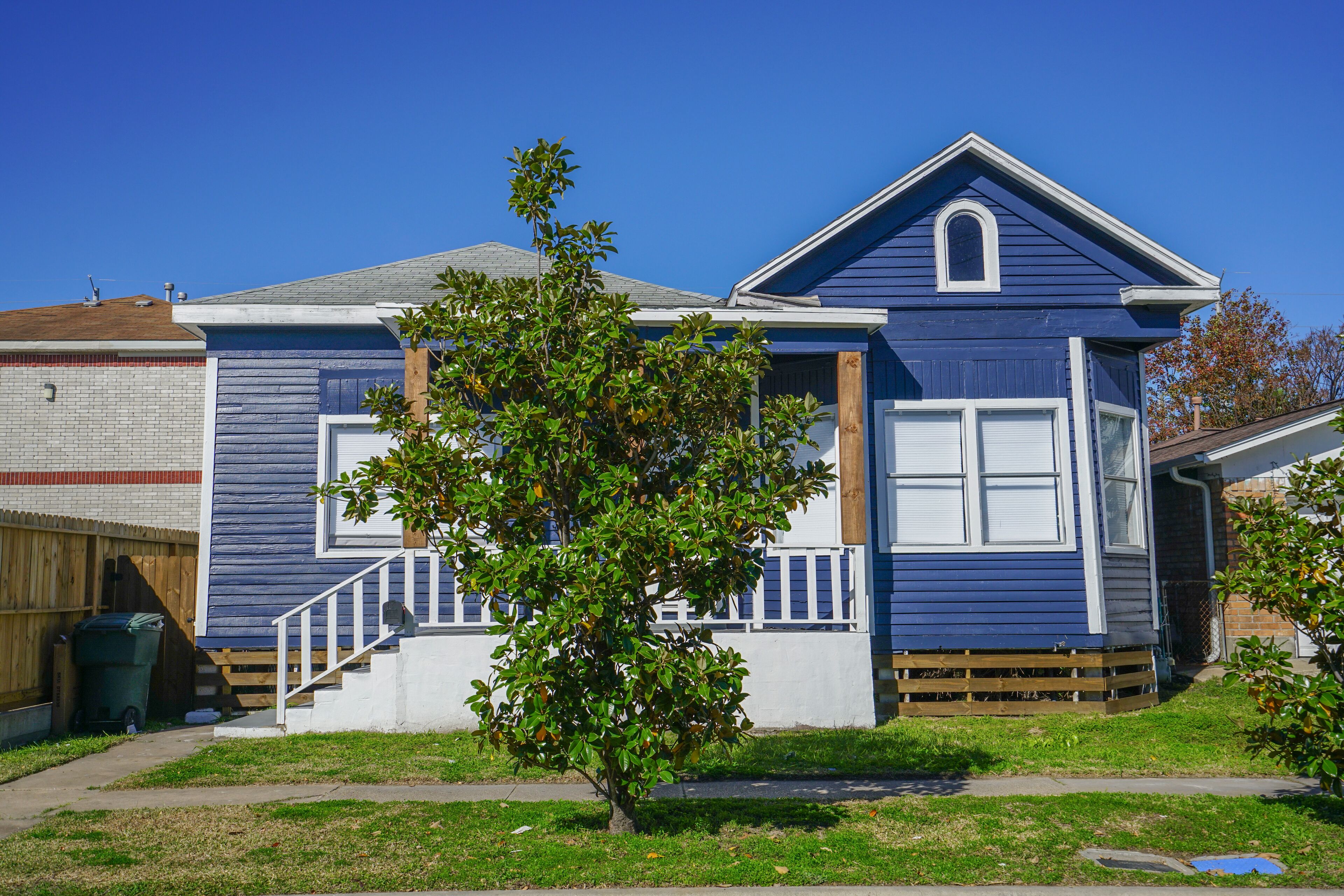 A two-story house with a front porch, a small tree in front, and a wooden fence in the background.