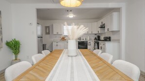 A dining area with a wooden table, white chairs, a vase with white feathers, and a kitchen in the background.