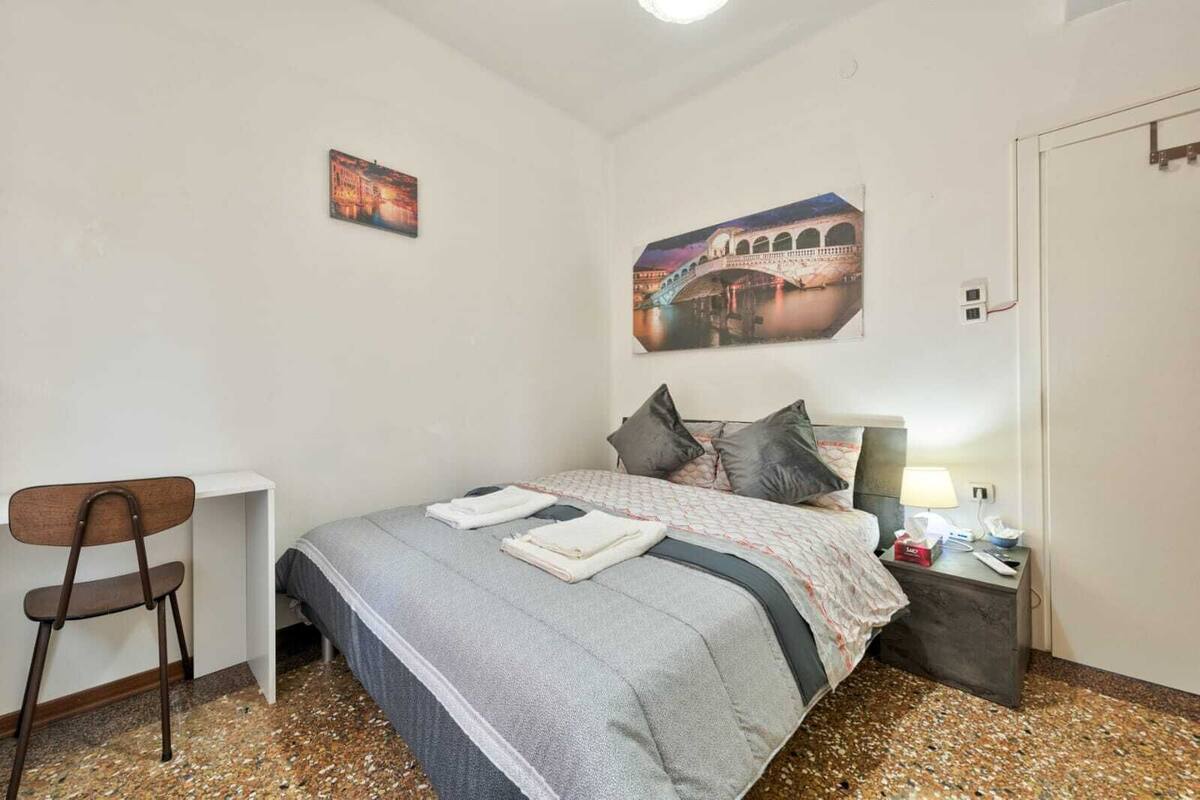 Un dormitorio ordenado con una cama, una silla, una mesita de noche con lámpara y dos cuadros colgados en la pared.