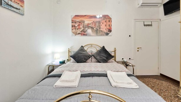 Un dormitorio ordenado con cama, almohadas, una mesita de noche, una lámpara y un cuadro con una vista urbana en la pared.