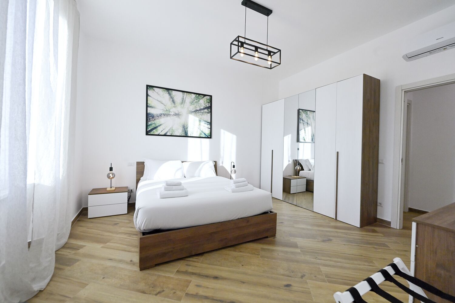 Quarto moderno com cama de madeira, roupa de cama branca, mesa de cabeceira com abajur, um espelho grande e um quadro na parede.