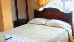 Una cama bien hecha con una colcha blanca y almohadas, una mesita de noche con una lámpara y una ventana con cortinas.